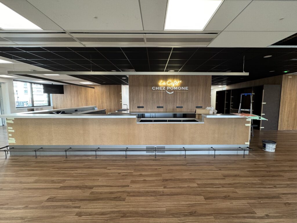 Vue d'ensemble de l'aménagement de l'espace "La Cafét Chez Pomone" : comptoir texturé, sol bois et enseigne lumineuse néon blanche sur mur bois zébré.
