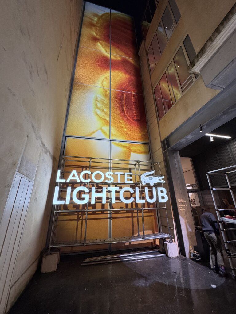 Installation verticale de l'enseigne lumineuse LACOSTE LIGHTCLUB à Paris avec bâche rétroéclairée grand format et structure métallique.