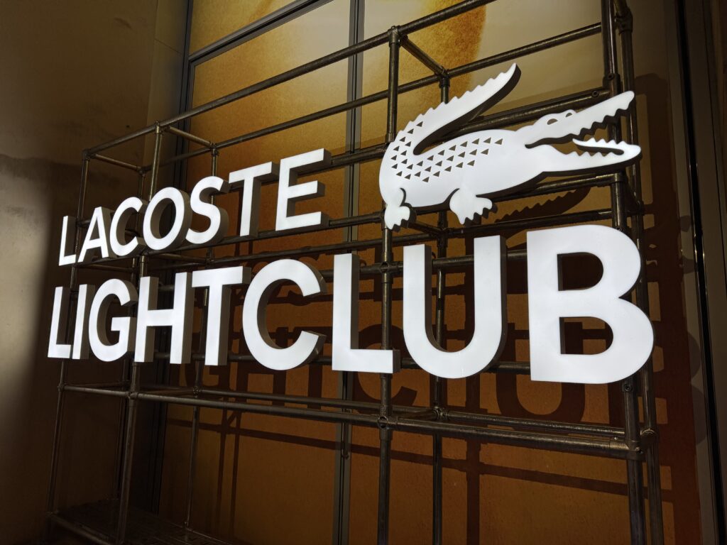 Gros plan de l'enseigne lumineuse LACOSTE LIGHTCLUB à Paris, avec lettres effet néon LED blanc froid et logo crocodile sur structure industrielle.
