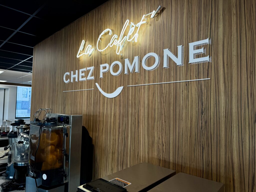 Enseigne lumineuse néon blanche et dorée "Le Café Chez Pomone" fixée sur un mur d'accent en bois, vue latérale.
